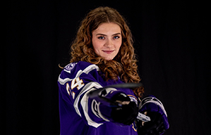 Woman holding a hockey stick towards the camera in front of a black curtain